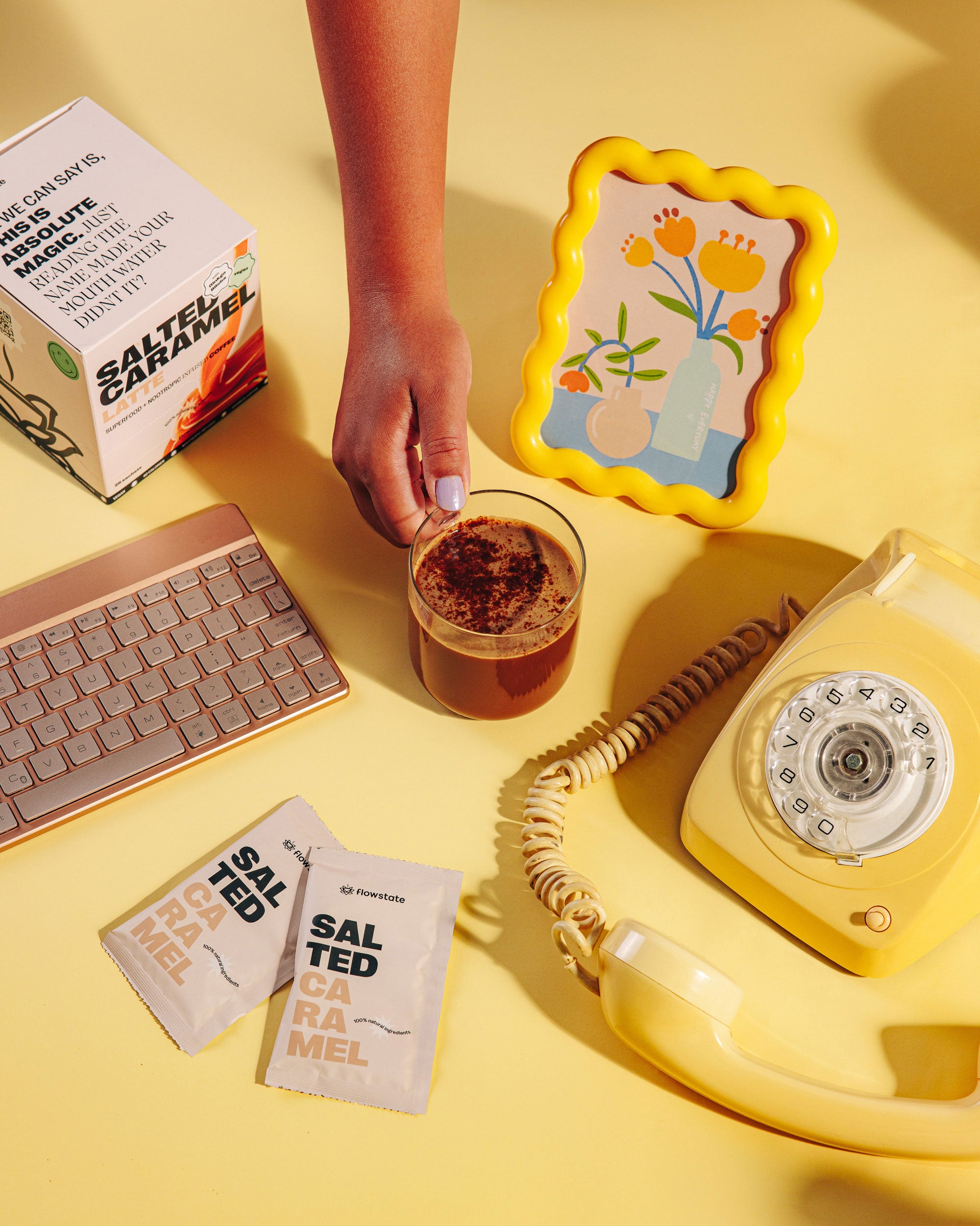 Hand grabbing cup of Flowstate coffee surrounded by keyboard, phone and Salted Caramel sachets